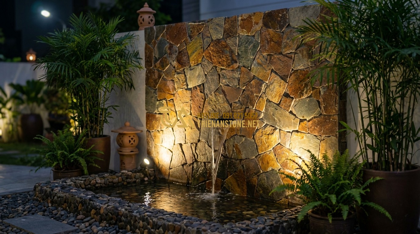 Tạo ảnh tiếng Anh: A modern garden feature wall made of dark grey slate irregular stones. A minimalist water fountain flows gently over the textured stones into a small pond with pebbles. Warm LED spotlights accentuate the irregular shapes and shadows of the stone wall at night. Zen garden atmosphere.