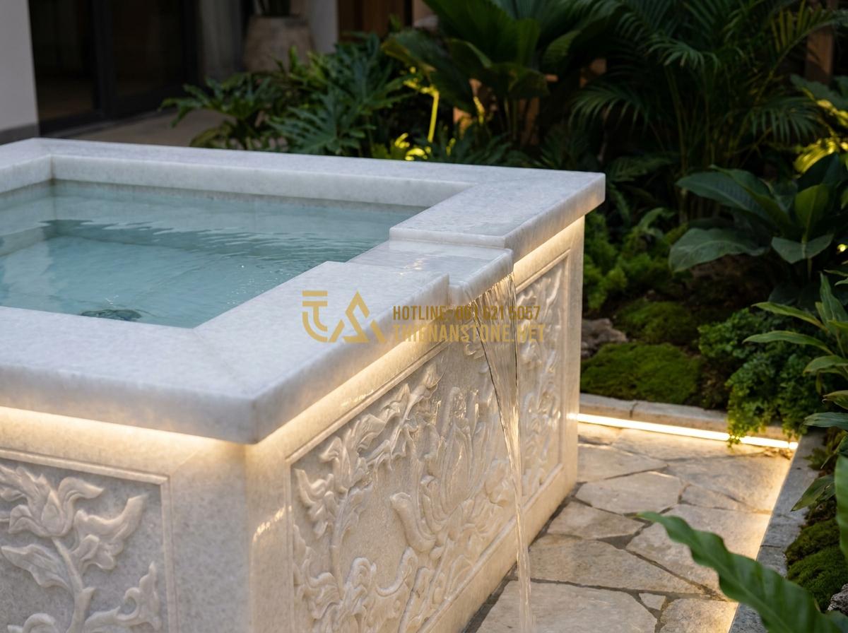 Tạo ảnh tiếng Anh: Luxurious close-up view of a modern square stone garden well made of polished natural basalt rock with intricate carvings, clear water gently flowing, integrated with soft LED stone lights illuminating the surrounding garden path at dusk, realistic professional photography style with warm ambient lighting and lush greenery background