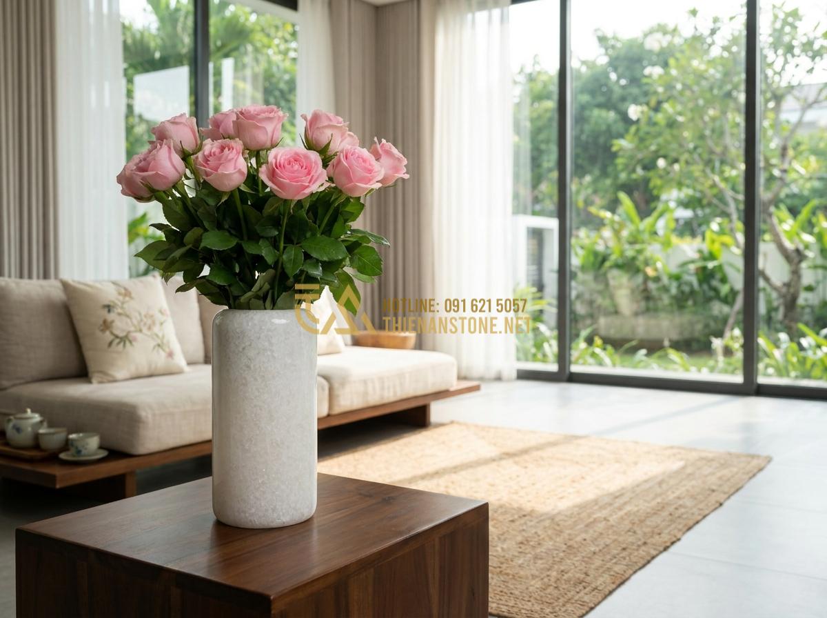 Tạo ảnh tiếng Anh: Professional high-resolution photograph of an elegant natural stone flower vase made from white marble with subtle gray veins, filled with fresh pink roses and green leaves, placed on a dark wooden side table in a bright minimalist living room with large windows, soft natural daylight streaming in, luxurious and serene atmosphere, photorealistic style, sharp details, 8k quality